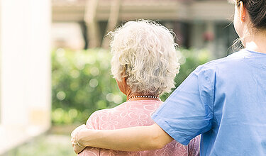 Junge Pflegerin geht mit einer älteren Dame im Garten spazieren und legt dabei ihren Arm auf ihre Schulter