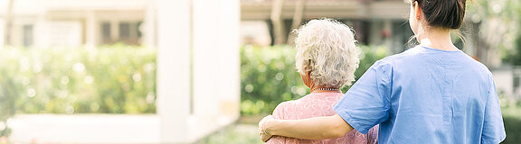 Junge Pflegerin geht mit einer älteren Dame im Garten spazieren und legt dabei ihren Arm auf ihre Schulter