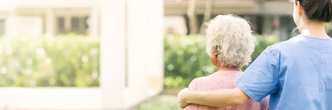 Junge Pflegerin geht mit einer älteren Dame im Garten spazieren und legt dabei ihren Arm auf ihre Schulter