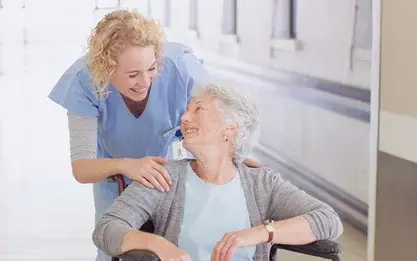 Ältere Dame, die in einem Rollstuhl sitzt, lächelt eine weibliche Pflegekraft an.