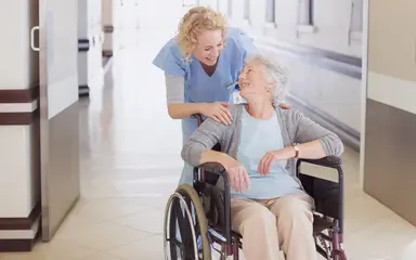 Ältere Dame, die in einem Rollstuhl sitzt, lächelt eine weibliche Pflegekraft an.