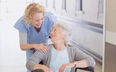 Ältere Dame, die in einem Rollstuhl sitzt, lächelt eine weibliche Pflegekraft an.