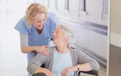 Ältere Dame, die in einem Rollstuhl sitzt, lächelt eine weibliche Pflegekraft an.