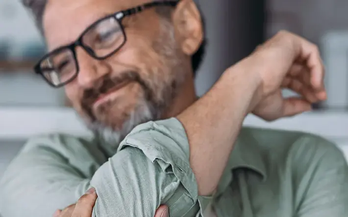 Ein Mann sitzt auf einem Sofa und hält den Arm. Dabei verzieht er aufgrund von Schmerzen das Gesicht.