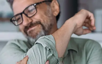 Ein Mann sitzt auf einem Sofa und hält den Arm. Dabei verzieht er aufgrund von Schmerzen das Gesicht.