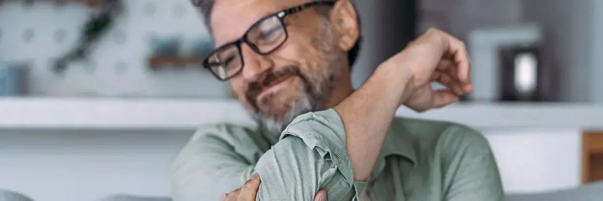 Ein Mann sitzt auf einem Sofa und hält den Arm. Dabei verzieht er aufgrund von Schmerzen das Gesicht.