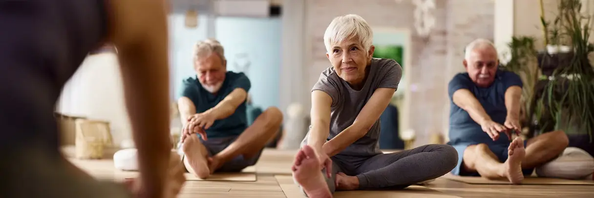 Ältere Menschen nehmen an einem Sportkurs teil. Auf dem Bild sind drei Personen mit grauen Haaren zu sehen, die Dehnungsübungen absolvieren.