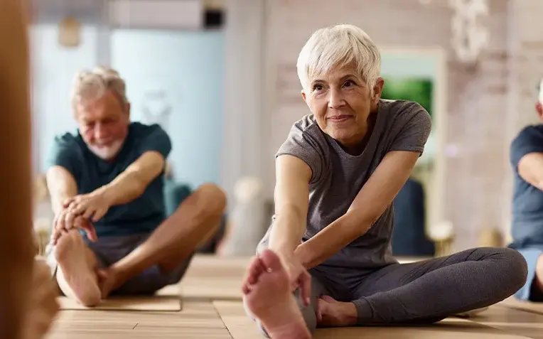 Ältere Menschen nehmen an einem Sportkurs teil. Auf dem Bild sind drei Personen mit grauen Haaren zu sehen, die Dehnungsübungen absolvieren.