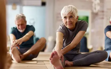Ältere Menschen nehmen an einem Sportkurs teil. Auf dem Bild sind drei Personen mit grauen Haaren zu sehen, die Dehnungsübungen absolvieren.
