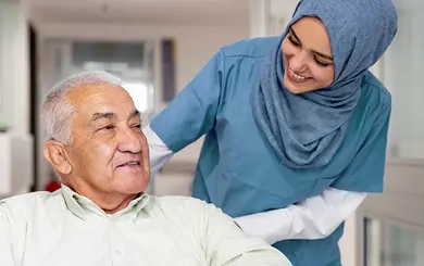 Pflegekraft mit Kopftuch im Gespräch mit einem älteren Mann in einer Pflegeeinrichtung.