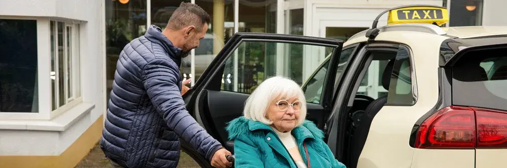 Ältere Frau im Rollstuhl vor Taxi, ein Mann hilft ihr beim Einsteigen, Szene vor einem Gebäude.
