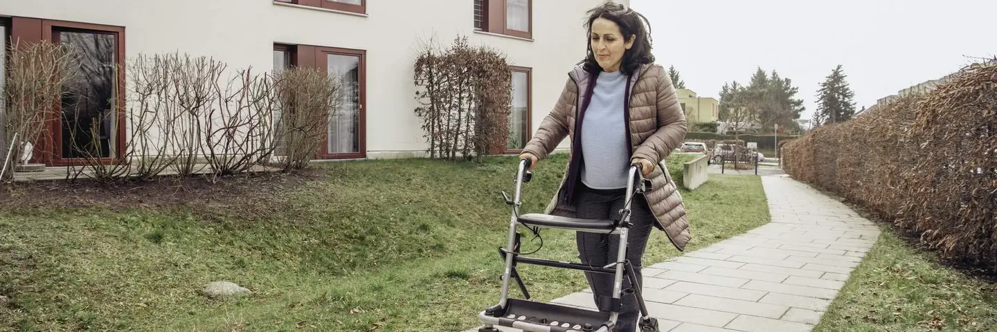 Frau Geht mit Rollator einen hellen gepflasterten Weg vor einem zweistöckigen Gebäude entlang