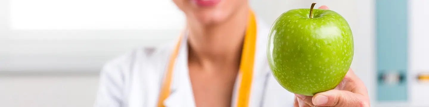 Junge Frau mit weißen Kittel und Maßband um ihren Hals, hält einen grünen Apfel 