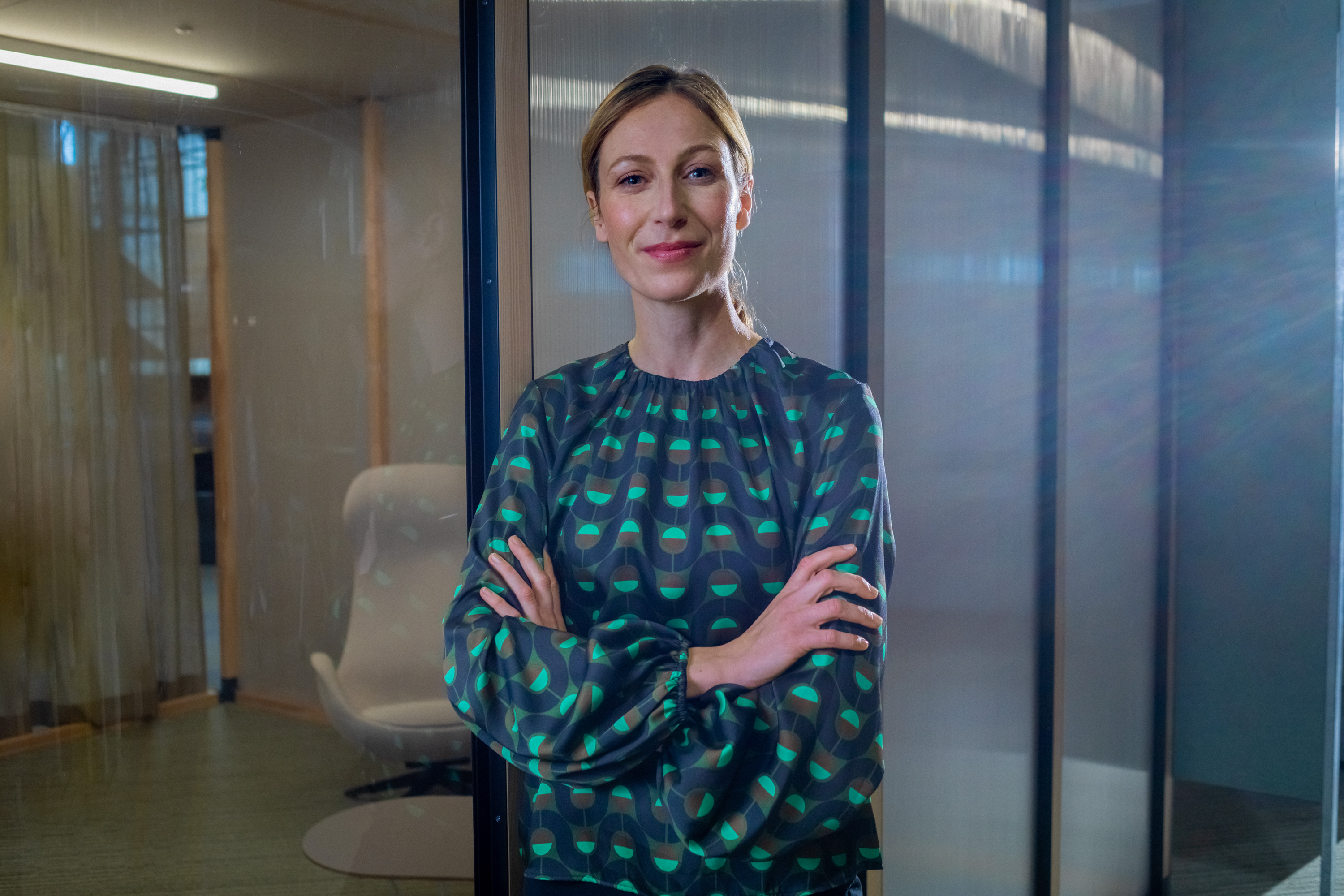 Eine berufserfahrene Frau steht mit verschränkten Armen vor einem verglasten Raum.