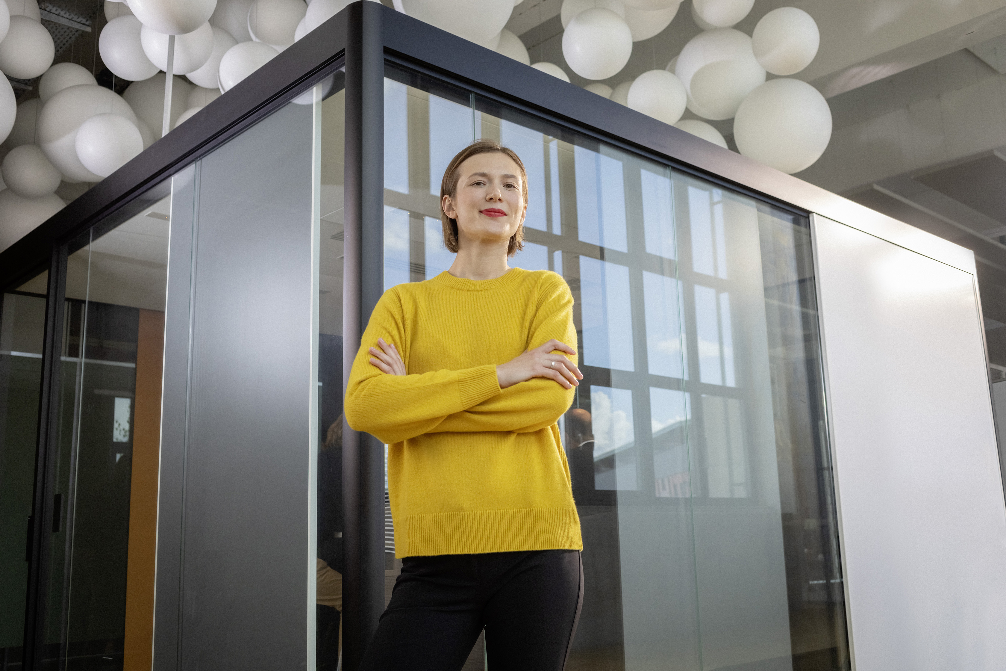 Eine berufserfahrene Frau steht mit verschränkten Armen vor einem verglasten Besprechungsraum.