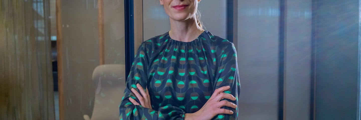 Eine berufserfahrene Frau steht mit verschränkten Armen vor einem verglasten Raum.