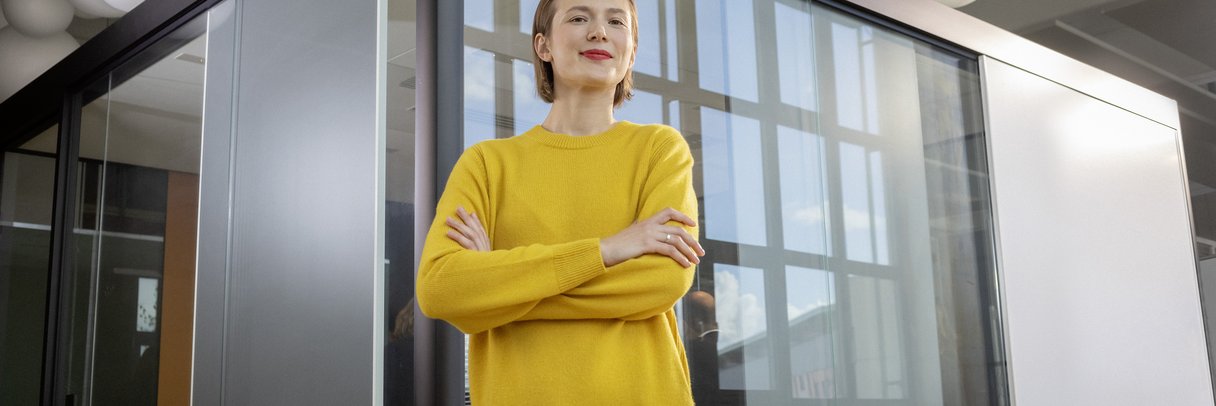 Eine berufserfahrene Frau steht mit verschränkten Armen vor einem verglasten Besprechungsraum.