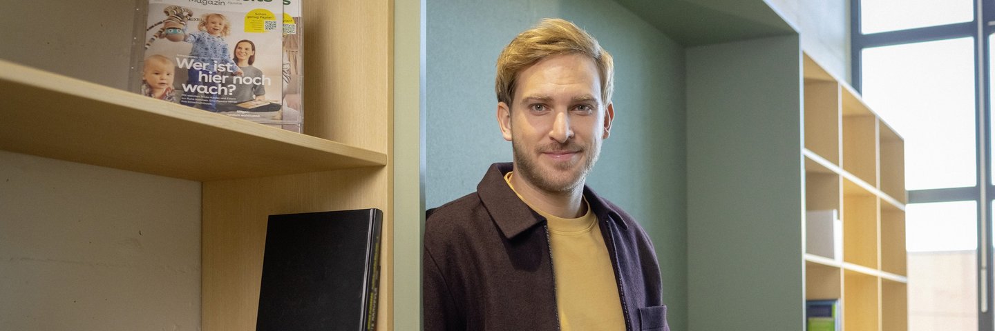Ein junger Berufserfahrener lehnt an einem Bücherregal.