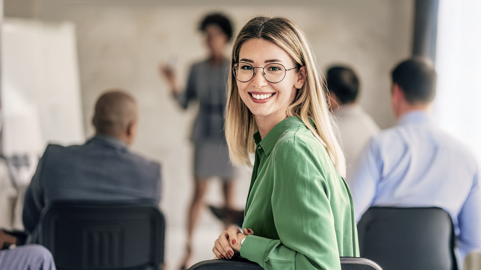 Lächelnde Frau in grünem Blazer sitzt in einem Seminarraum und blickt freundlich in die Kamera, im Hintergrund findet eine Präsentation statt.