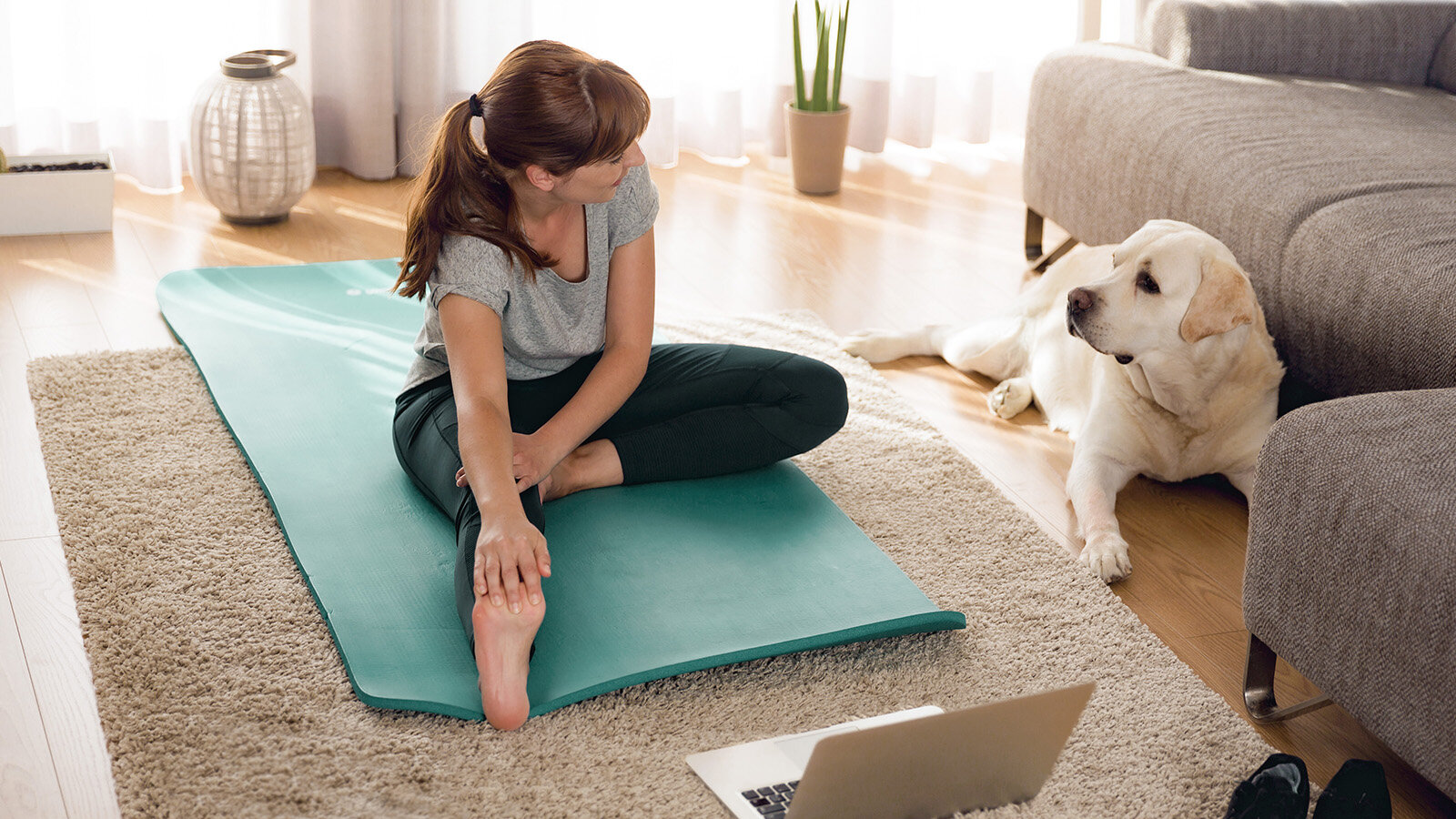 Eine junge Frau dehnt sich auf ihrer Yogamatte und schaut ihren Hund an.