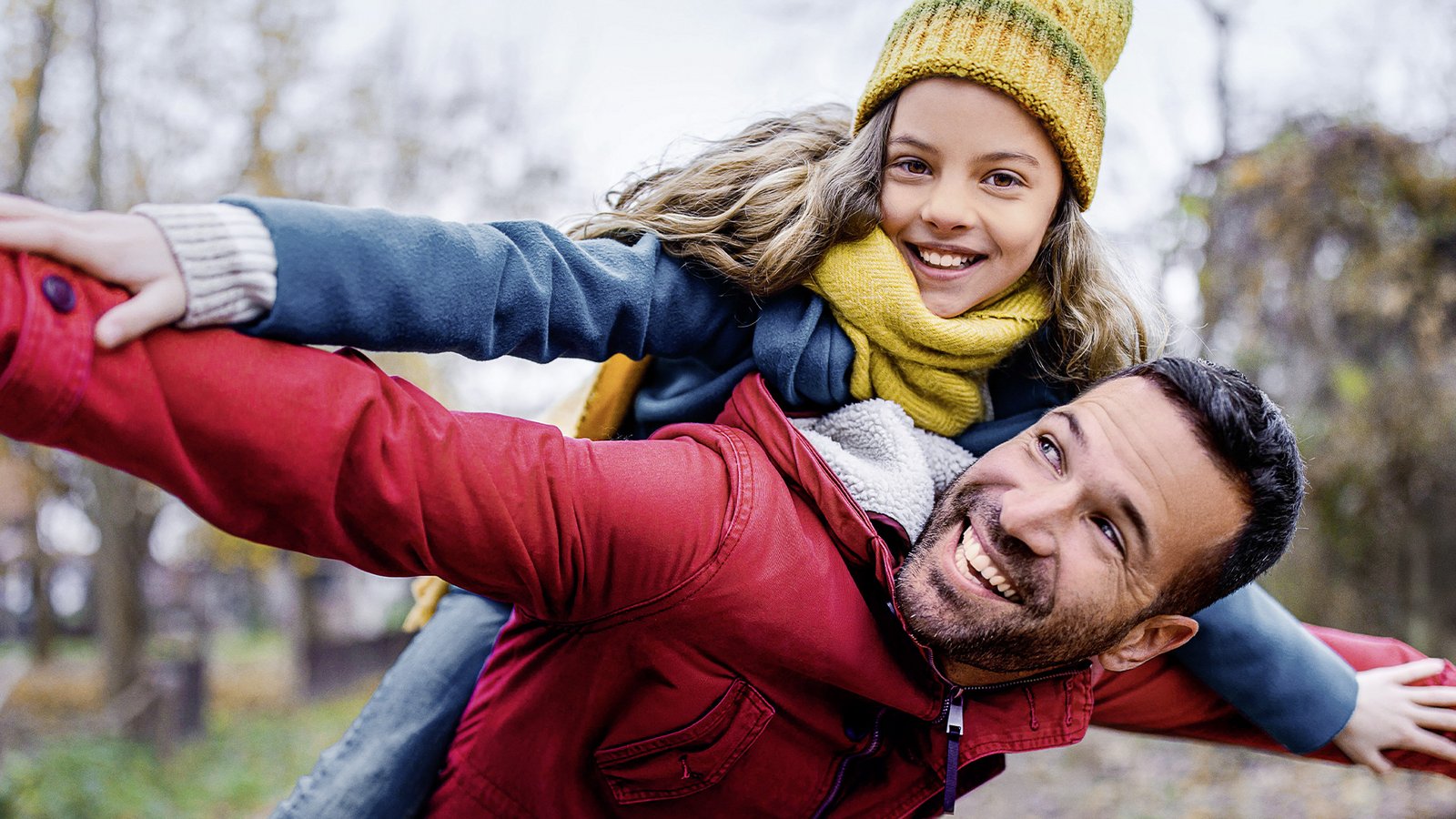 Vater trägt sein lachendes Kind huckepack im herbstlichen Park; beide breiten spielerisch die Arme aus wie beim Fliegen.
