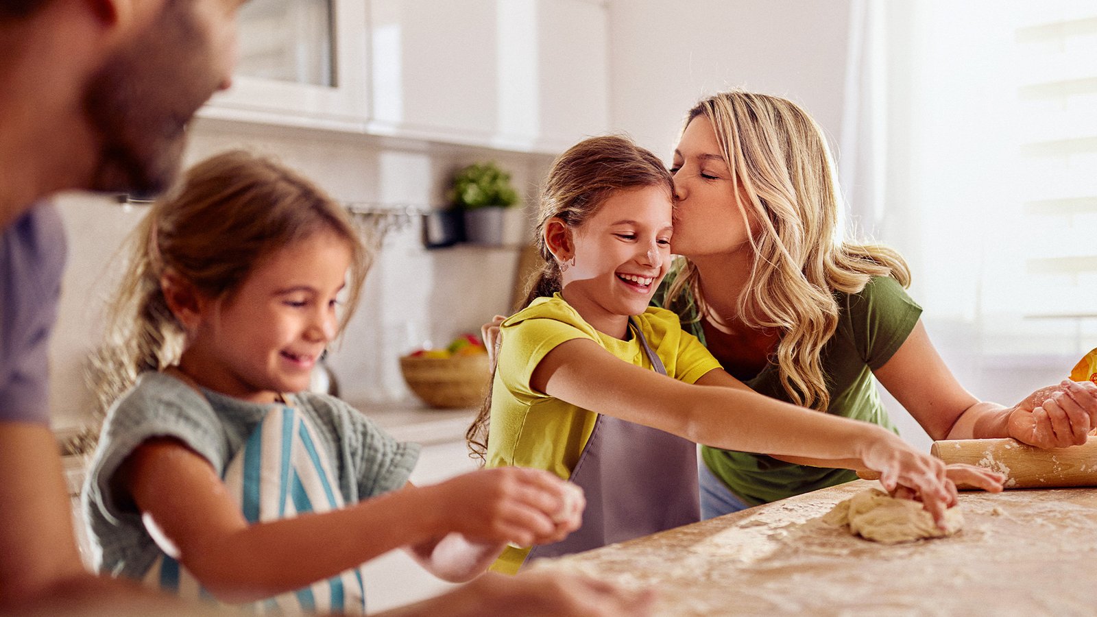 Familie backt gemeinsam in der Küche, Kinder kneten Teig, eine erwachsene Person gibt einem Kind einen liebevollen Kuss auf den Kopf.