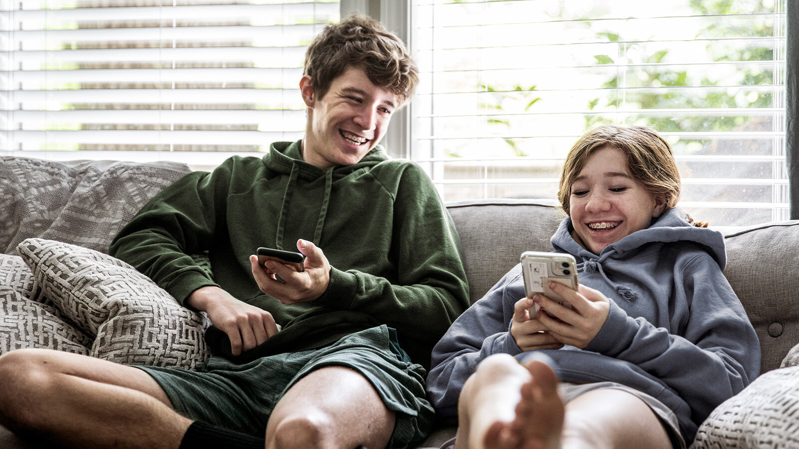 Junge und Mädchen sitzen mit Smartphones in den Händen auf einer Couch und lachen