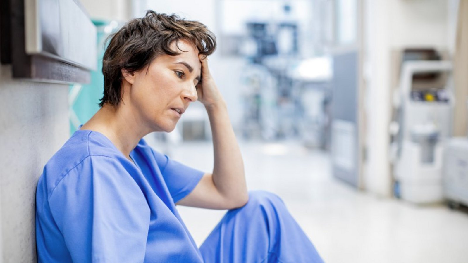 Pflegekraft in Arbeitskleidung sitzt erschöpft auf dem Boden eines Klinikflurs und stützt den Kopf mit der Hand.