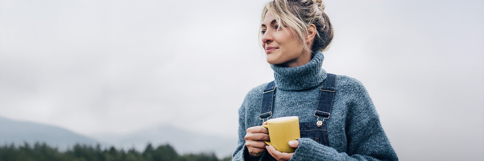 Frau in warmer Outdoor-Kleidung hält eine gelbe Tasse und blickt entspannt in die Ferne vor einem nebligen Berg- und Waldpanorama.