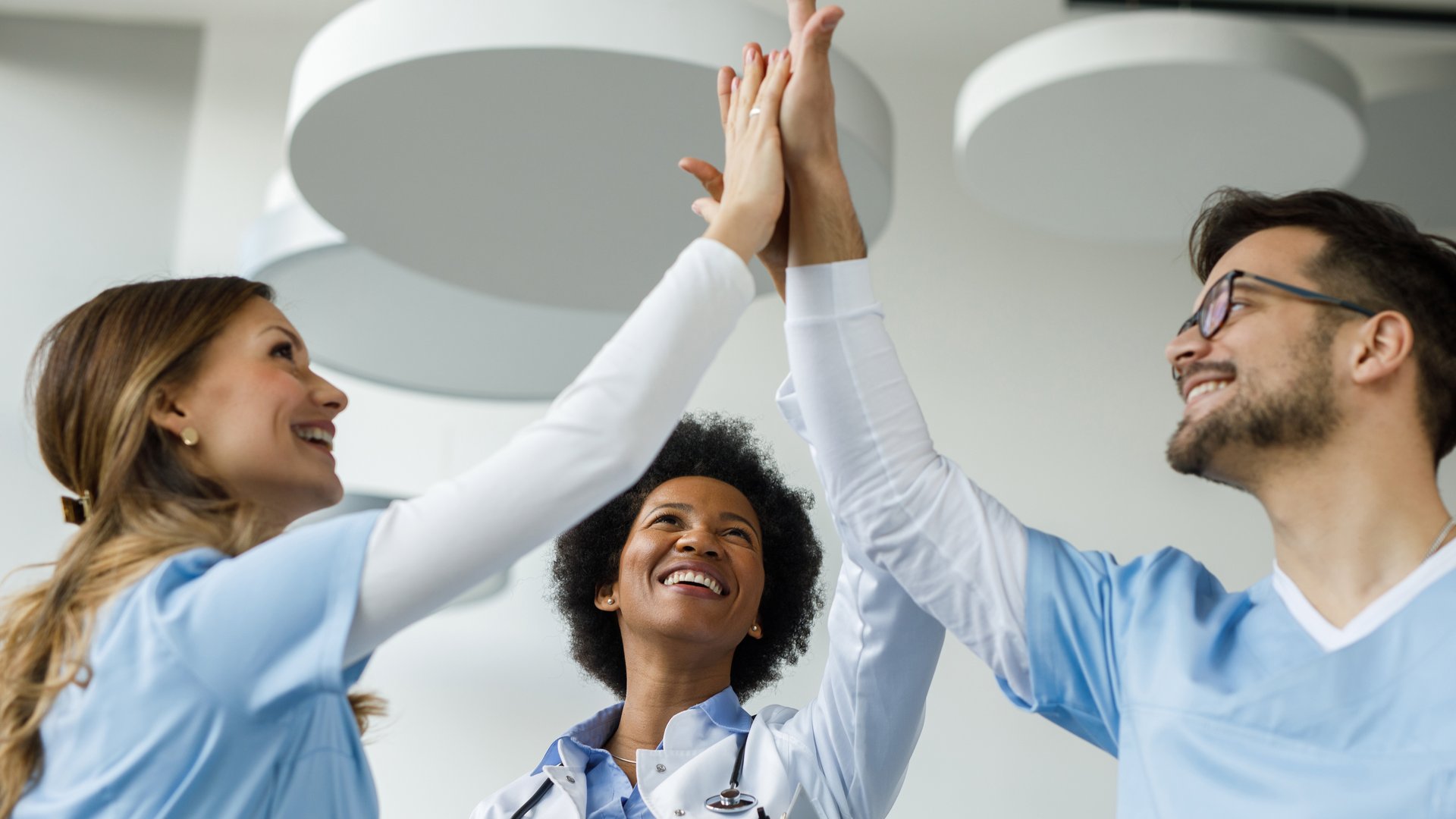 Drei Mitarbeitende im Gesundheitswesen stehen zusammen und geben sich lächelnd ein High Five. Eine Person trägt einen weißen Kittel und ein Stethoskop, die beiden anderen tragen hellblaue Arbeitskleidung.