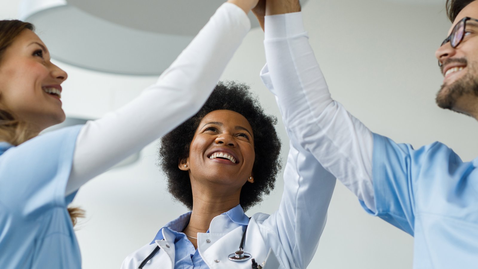 Drei Mitarbeitende im Gesundheitswesen stehen zusammen und geben sich lächelnd ein High Five. Eine Person trägt einen weißen Kittel und ein Stethoskop, die beiden anderen tragen hellblaue Arbeitskleidung.