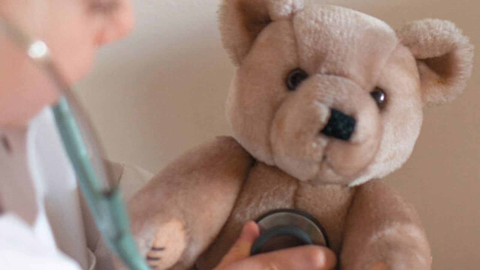 Teddy mit von Kinderhand gehaltenem Stethoskop auf der Brust, rechts daneben leicht verschwommenes Kindergesicht. 