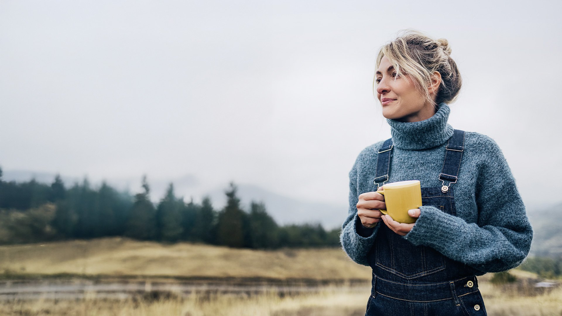 Frau in warmer Outdoor-Kleidung hält eine gelbe Tasse und blickt entspannt in die Ferne vor einem nebligen Berg- und Waldpanorama.