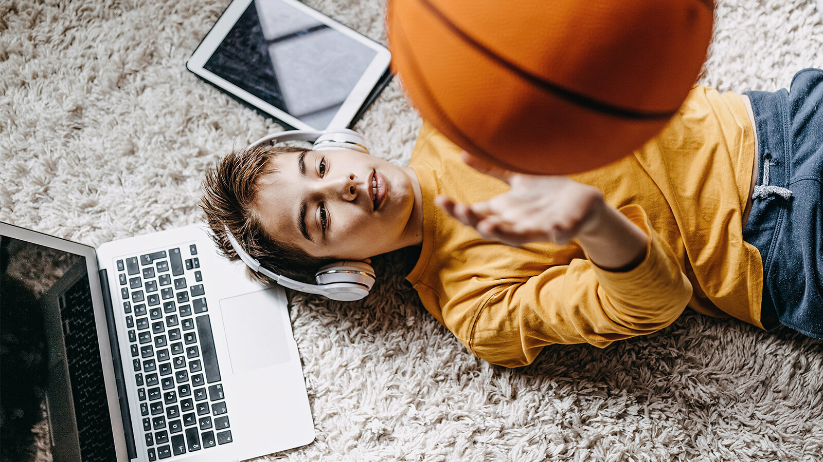 Junge mit Kopfhörern liegt auf einem Teppich und wirft einen Basketball in die Luft
