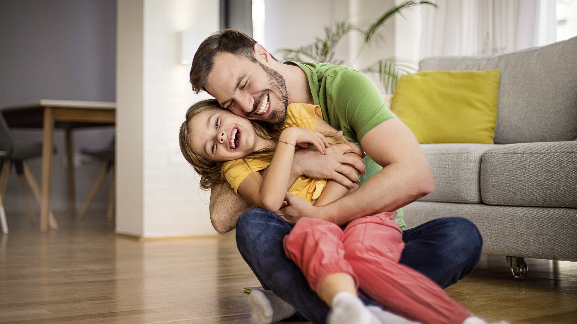 Vater sitzt auf dem Boden und umarmt lachend sein Kind im Wohnzimmer; beide strahlen Freude und Nähe aus.