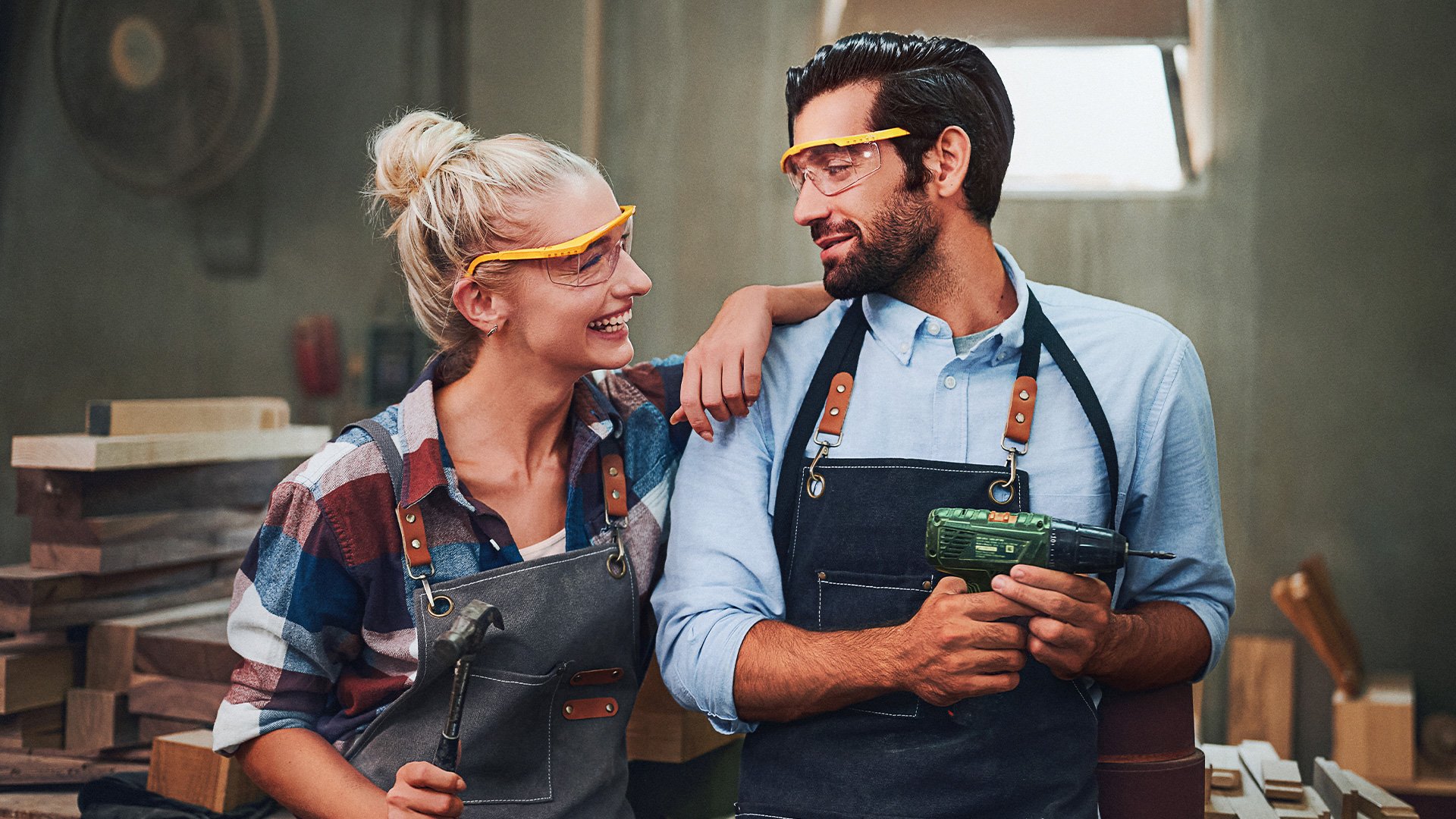 Zwei Personen in Arbeitskleidung und Schutzbrillen stehen lachend in einer Werkstatt und halten Werkzeug in den Händen.