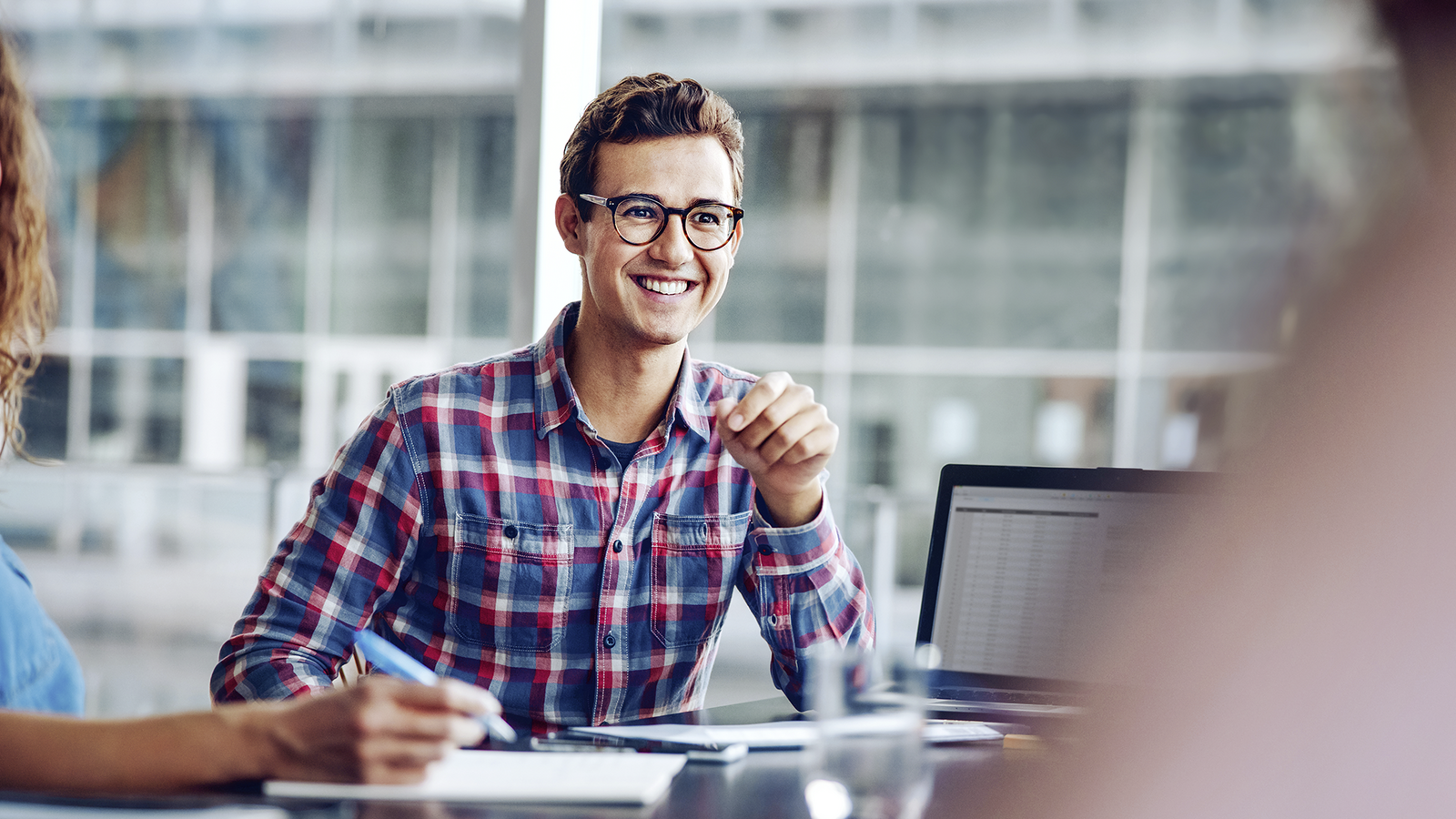Junger Mann mit Brille im karierten Hemd sitzt in einem modernen Büro an einem Tisch mit Laptop und lächelt während eines Gesprächs.