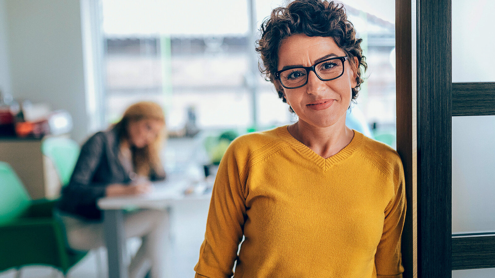 Bild zeigt Frau in gelbem Pullover, die im Büro an einen Türrahmen gelehnt, lächelnd in die Kamera schaut, freut sich über das Angebot Mehrleistungen.