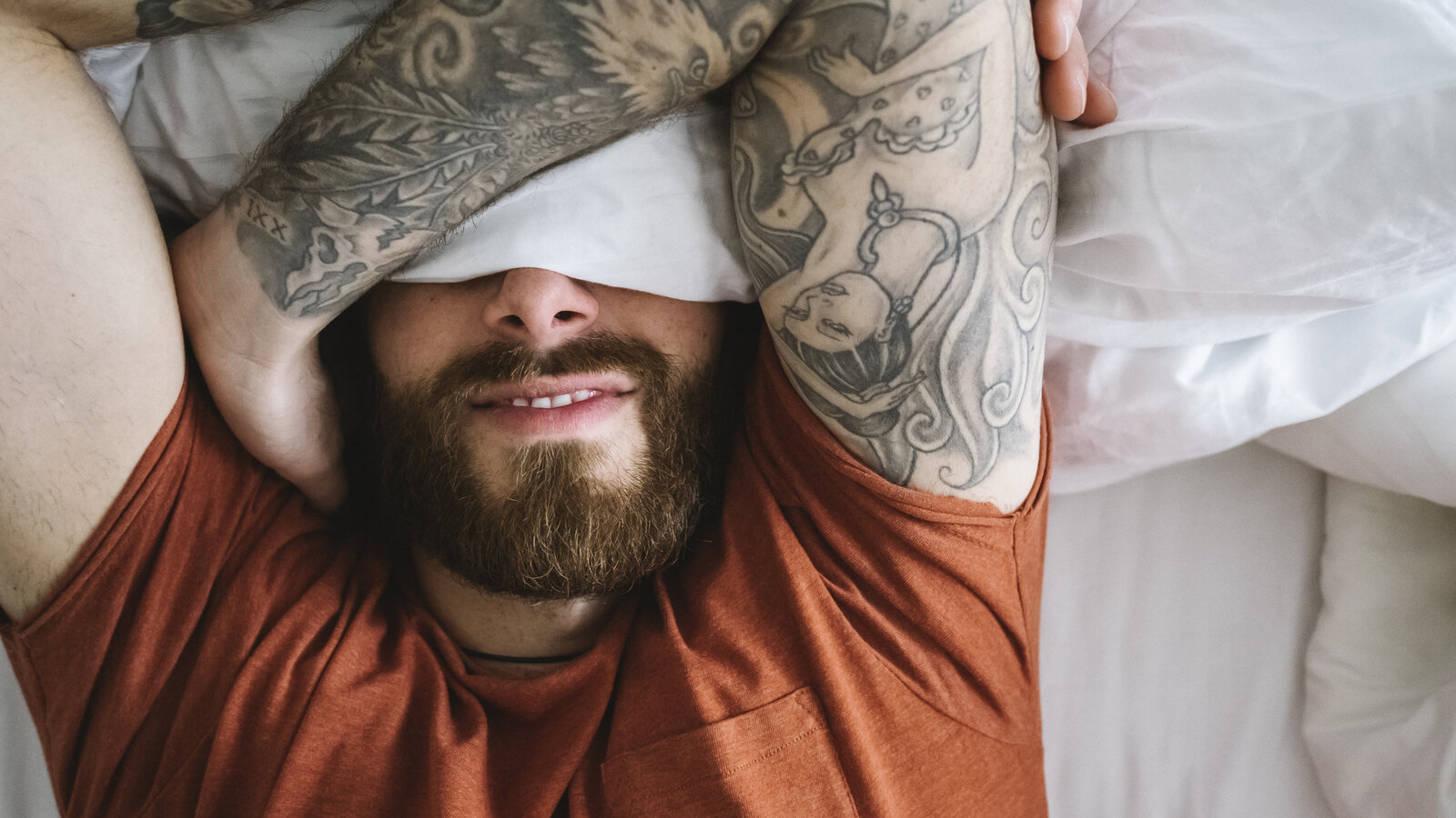 Mann mit Bart und taetowierten Armen liegt im Bett und verschraenkt die Arme vor den Augen.
