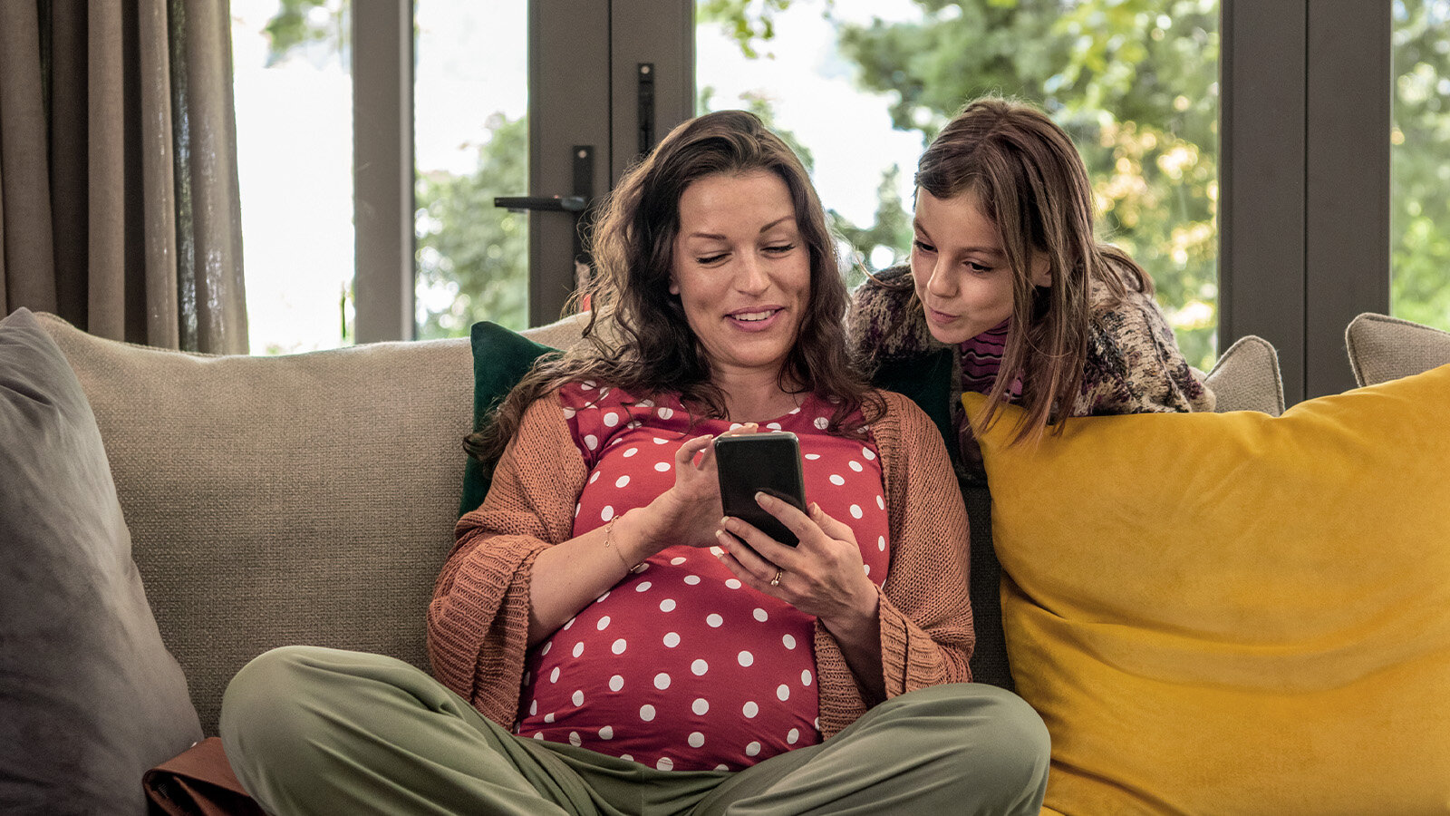 Eine Frau sitzt auf den Sofa mit dem Handy und ihre Tochter schaut ihr dabei zu 