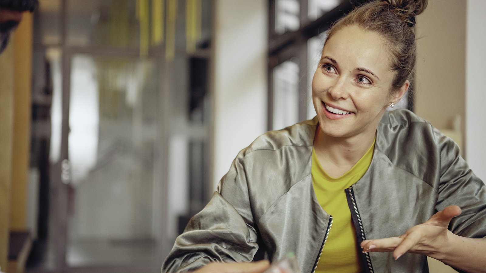 Frau sitzt an einem Tisch, lächelt und spricht gestikulierend mit einer anderen Person in einem modernen Innenraum.