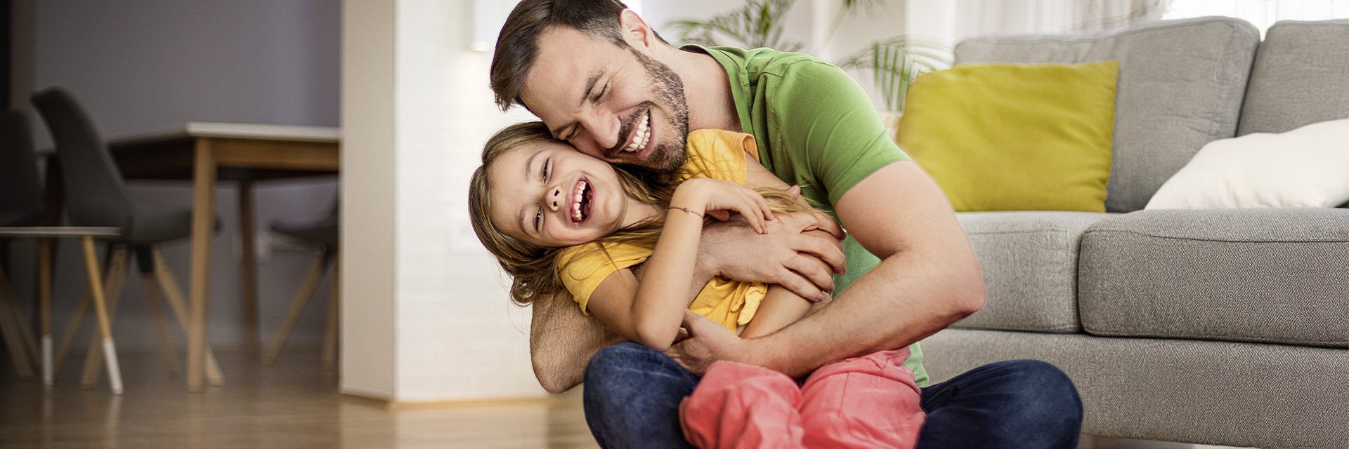 Vater sitzt auf dem Boden und umarmt lachend sein Kind im Wohnzimmer; beide strahlen Freude und Nähe aus.