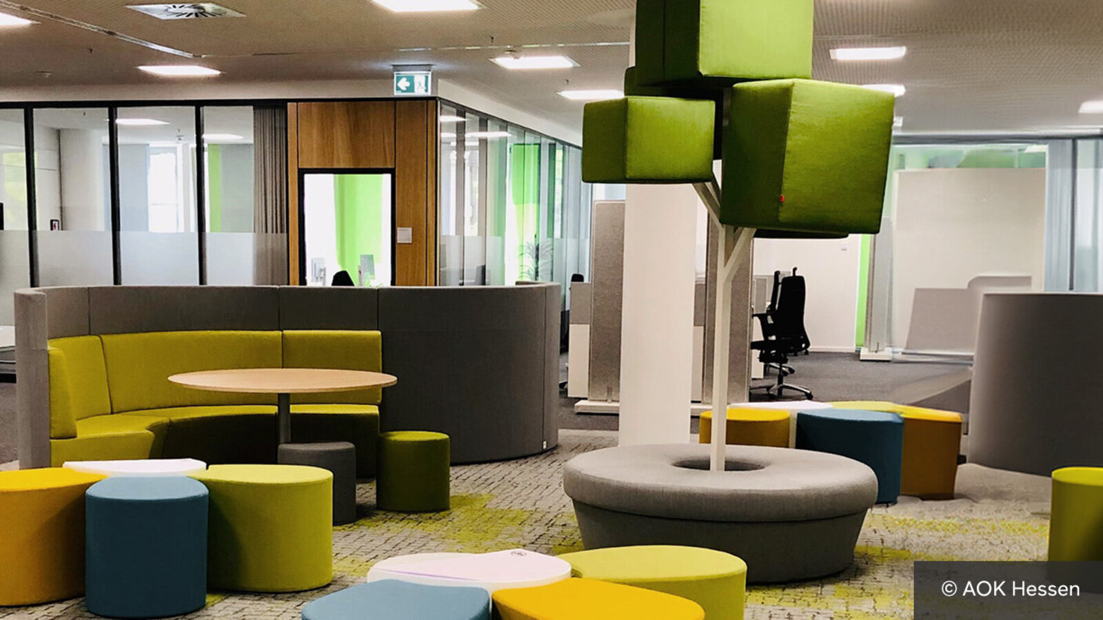 In der Mitte einer Büroetage, zwischen mehreren Büros, steht ein Dutzend bunter Hocker, ein gelbes Ecksofa und ein runder Holztisch. 