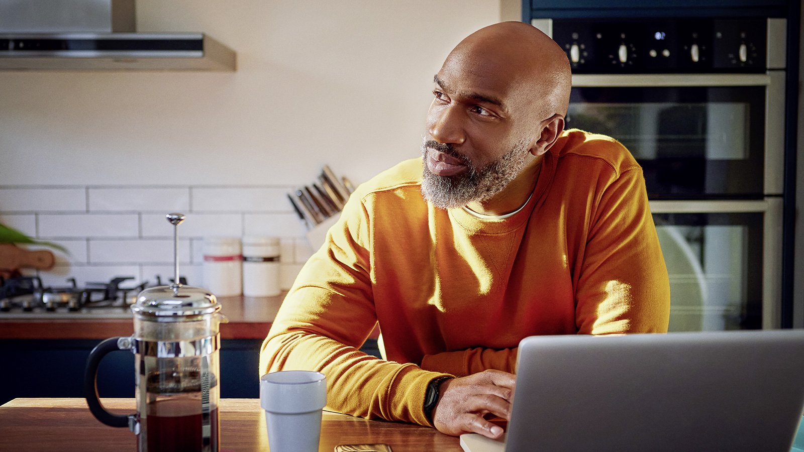 Mann sitzt am Küchentisch neben einer Kaffeekanne und einem Laptop, blickt nachdenklich zur Seite und genießt eine ruhige Momentpause.