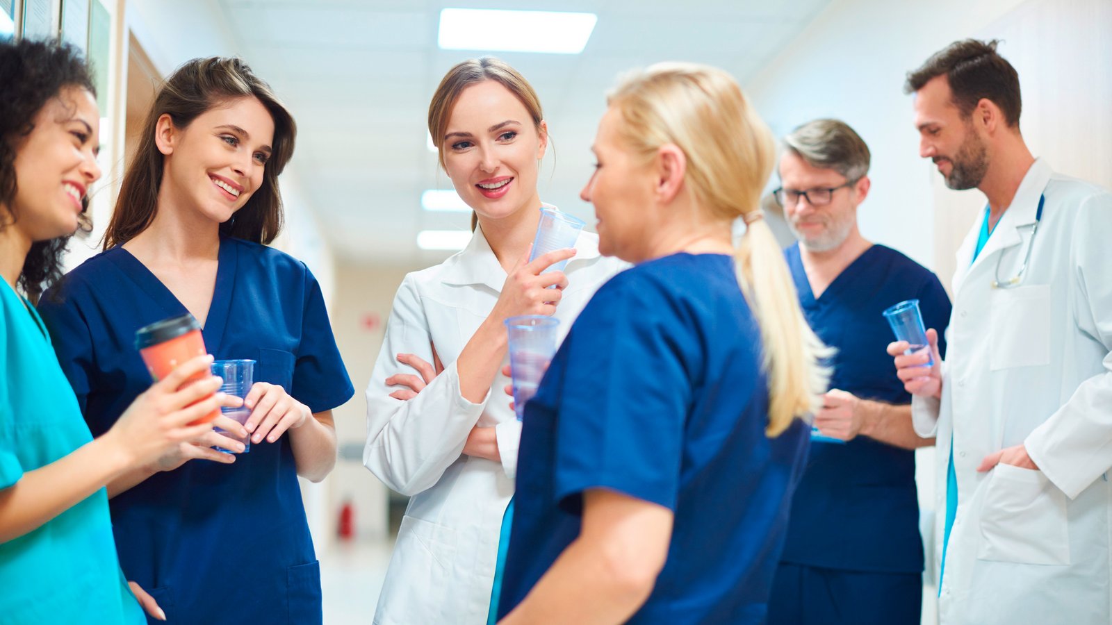 Mehrere Ärztinnen, Ärzte und Pflegekräfte stehen in einem Krankenhausflur zusammen und unterhalten sich während einer Pause. Einige halten Becher in der Hand und wirken entspannt.