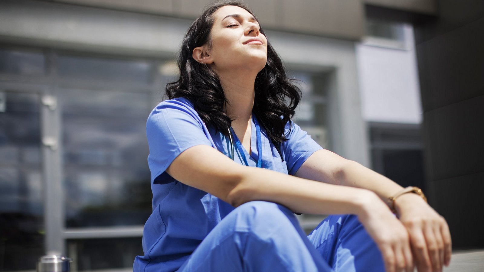 Eine weibliche Pflegekraft sitzt draußen auf einer Treppe und richtet den Kopf mit geschlossenen Augen in die Sonne.