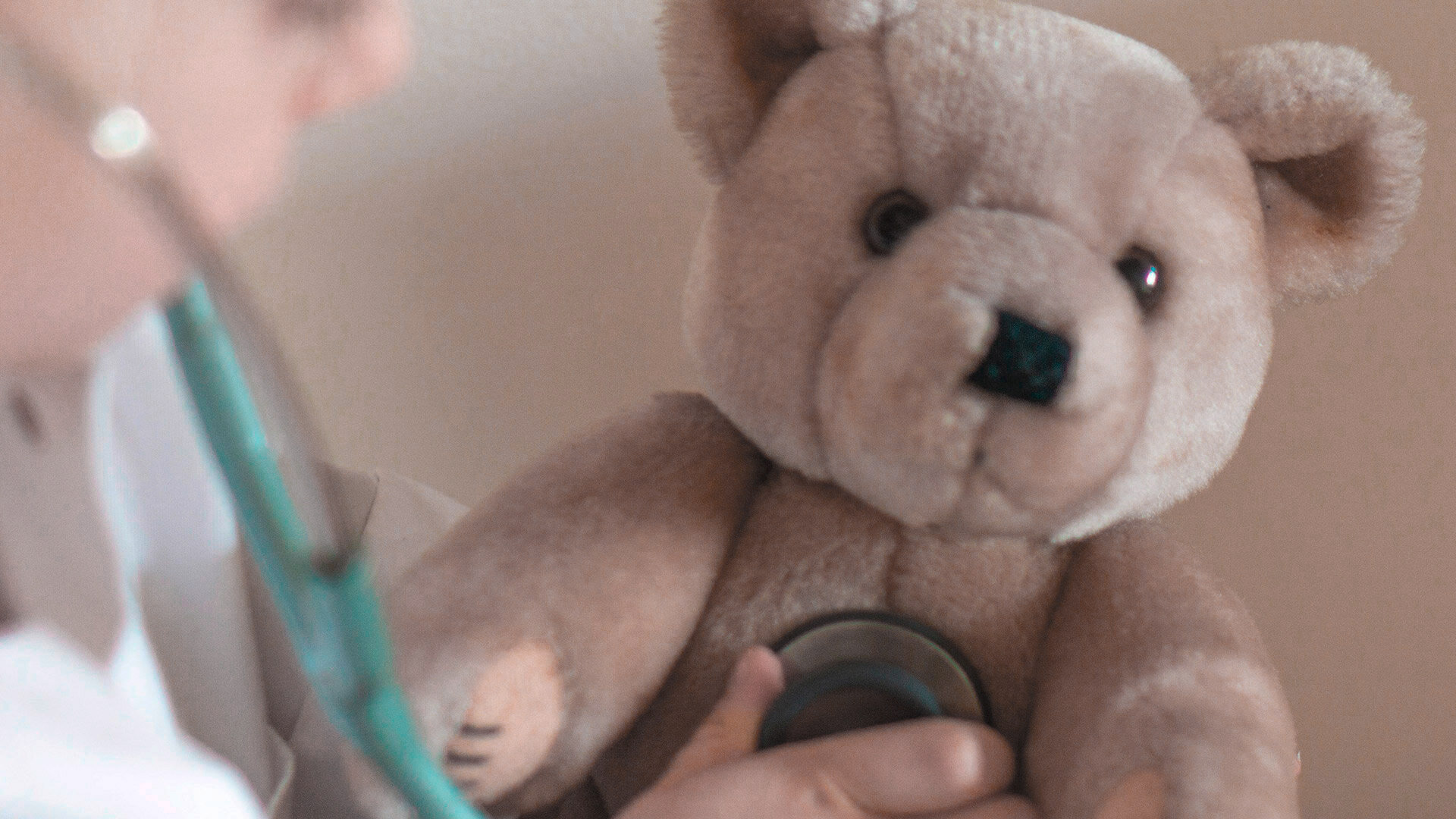 Teddy mit von Kinderhand gehaltenem Stethoskop auf der Brust, rechts daneben leicht verschwommenes Kindergesicht.