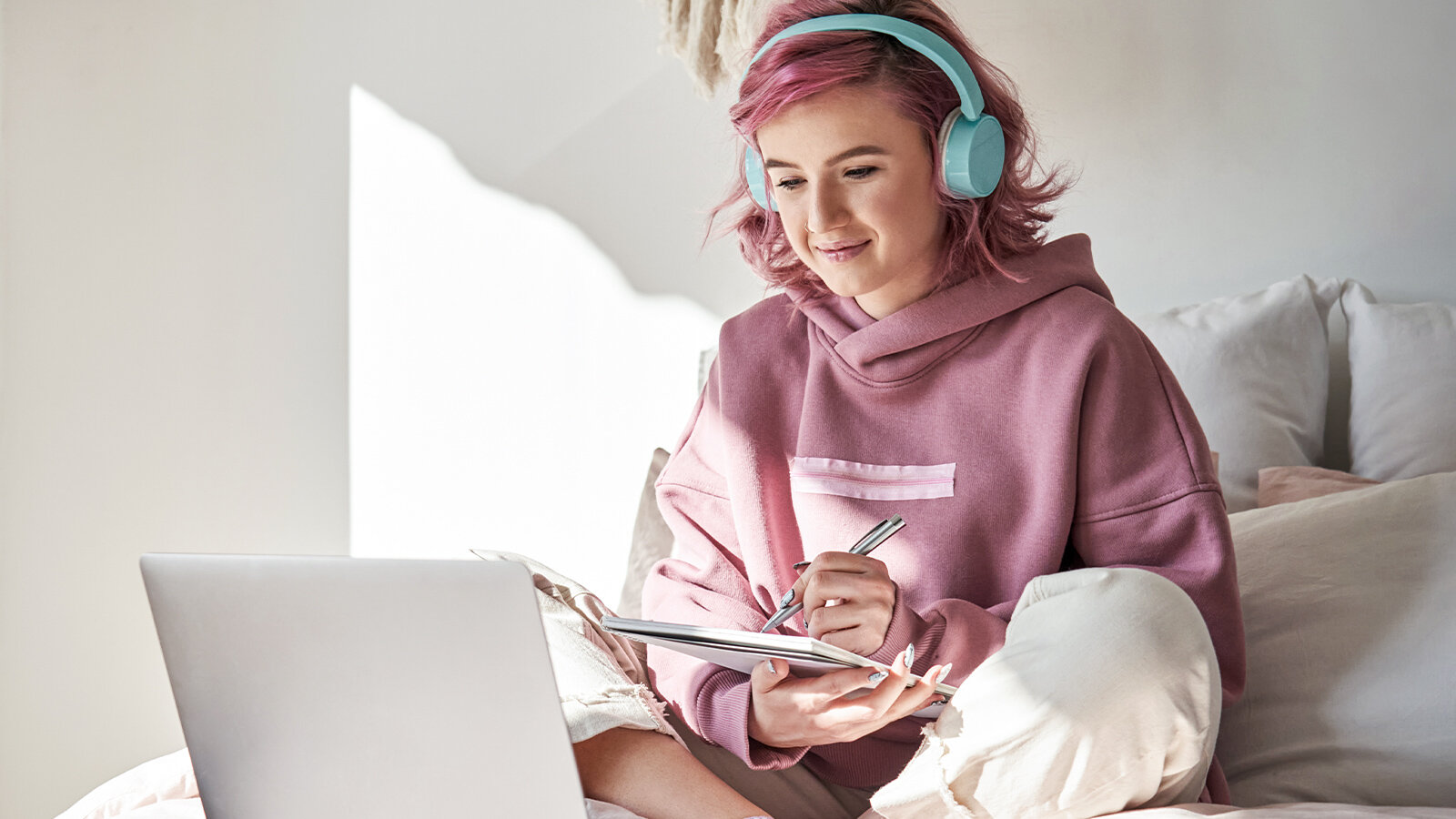 Eine junge Frau mit Kopfhörern sitzt mit einem Laptop vor sich auf dem Bett