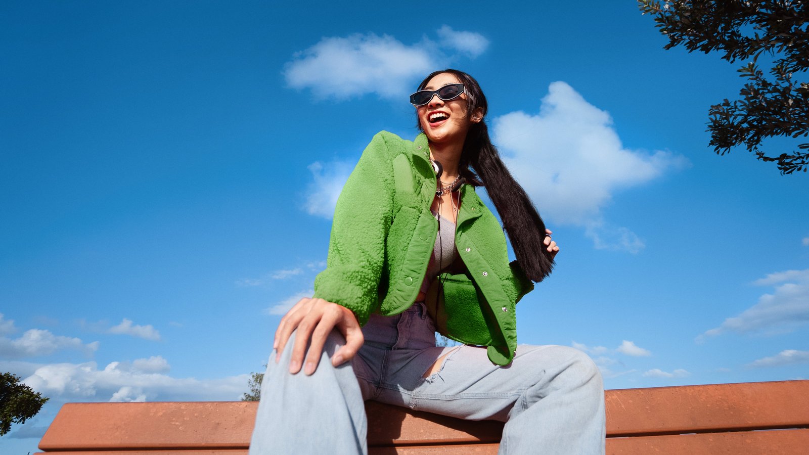 Junge Frau mit Sonnenbrille und grüner Jacke sitzt draußen auf einer Bank unter blauem Himmel und lacht.