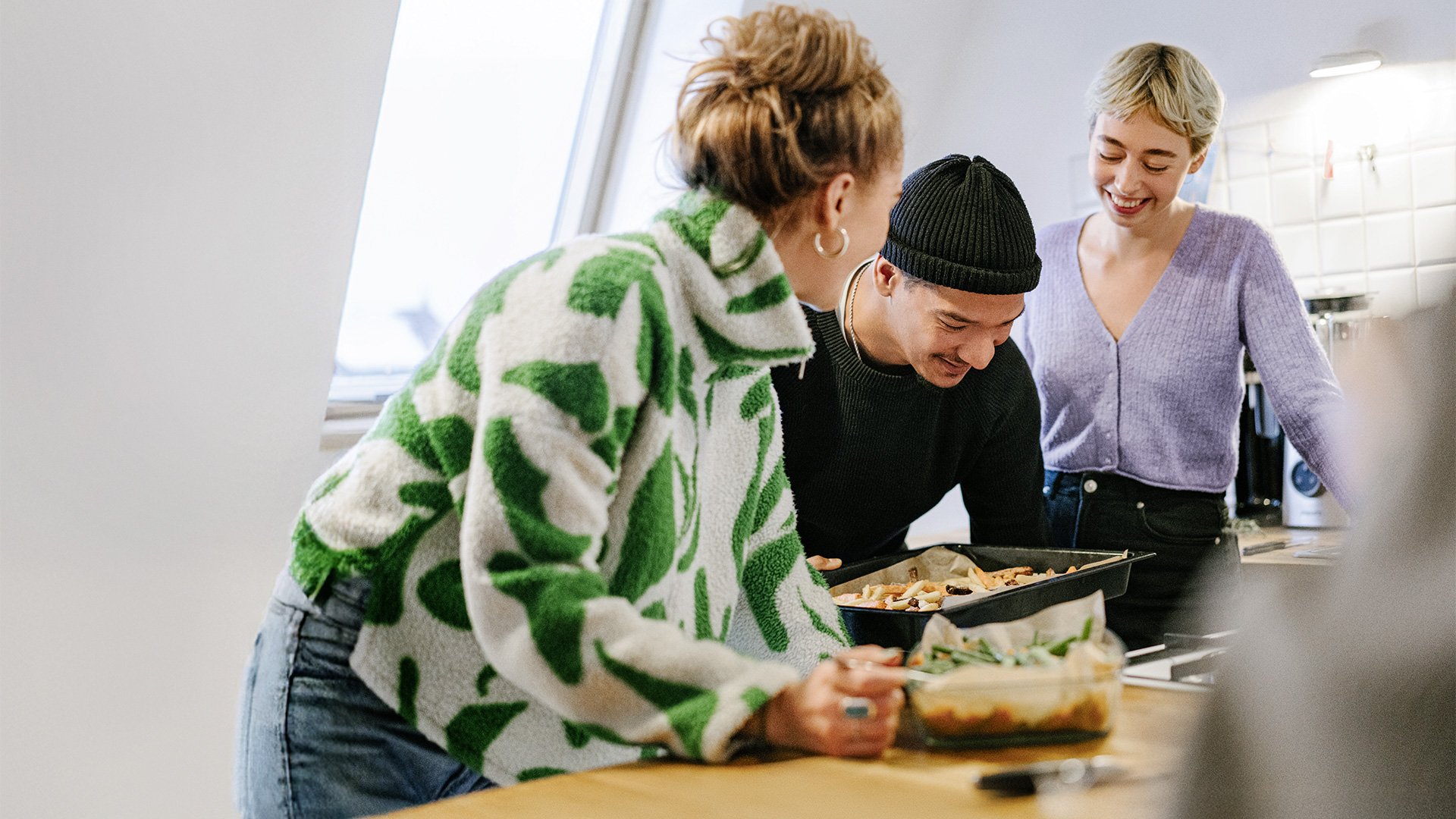 Drei junge Menschen kochen zusammen in einer Küche, lachen und betrachten ein Blech mit frisch zubereitetem Essen.